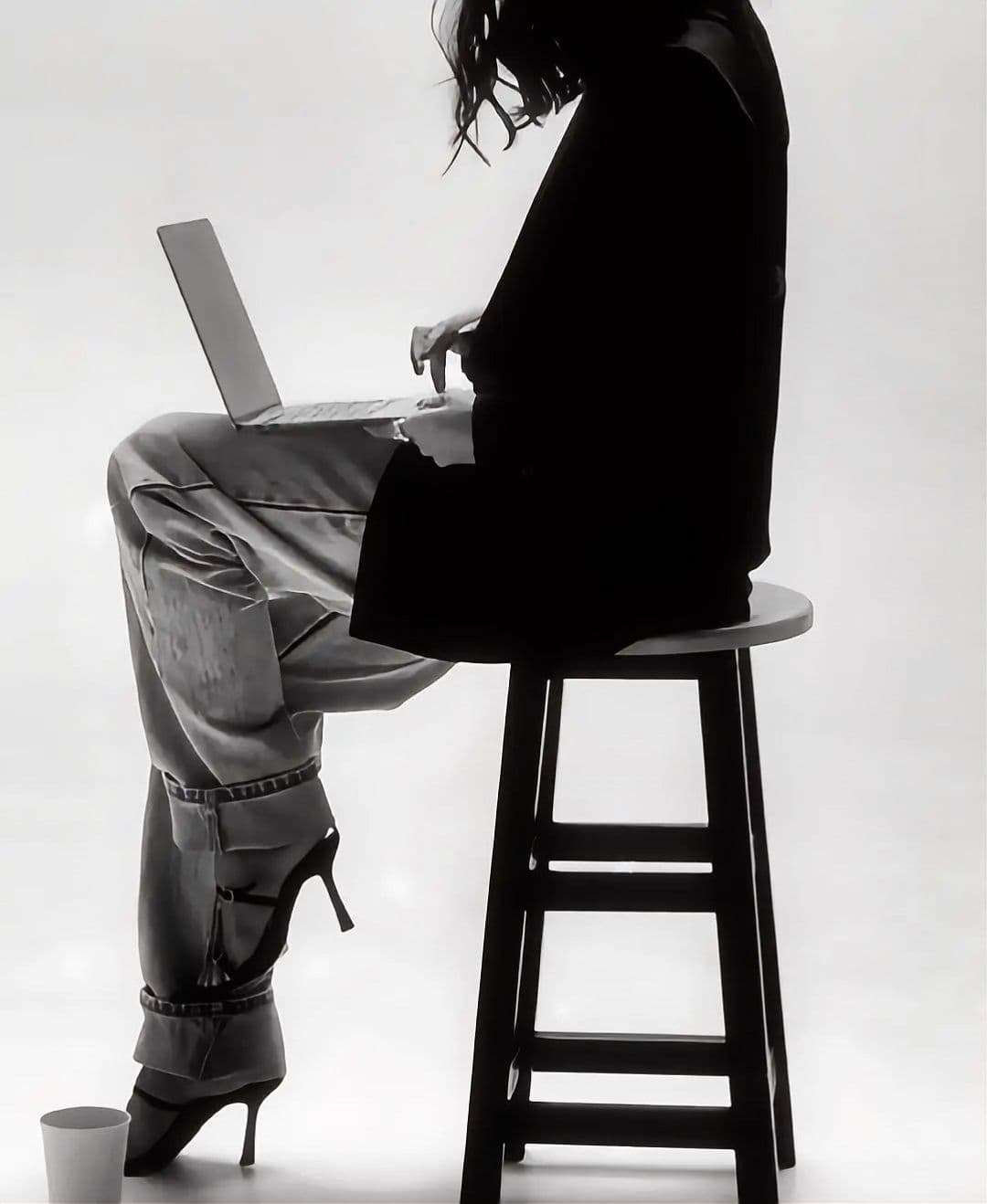Woman in loose pants and heels sits on a stool using a laptop.